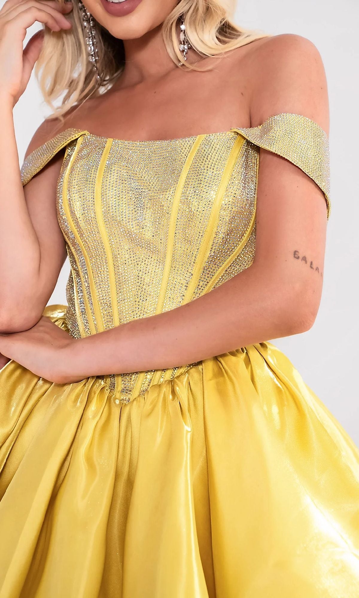 A woman wears the Portia and Scarlett PS24762 short homecoming dress, which features an off-shoulder yellow satin skirt and a glittery rhinestone corset bodice. Only her torso and part of her face are shown.